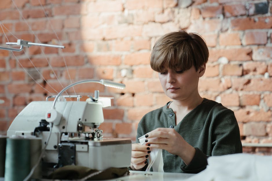 Female tailor focusing on sewing a fabric using a sewing machine in a brick walled workshop.