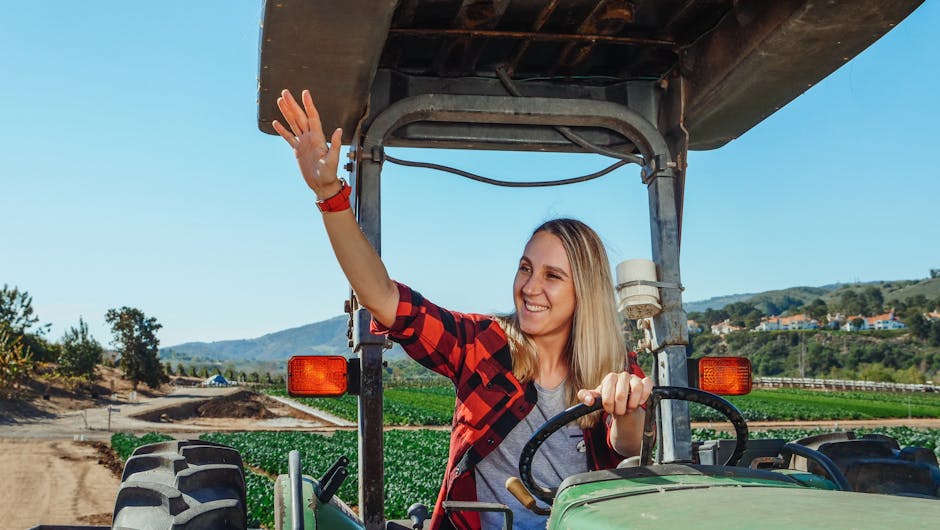 Woman waving while driving a tractor on a sunny day at a farm, showcasing rural lifestyle.